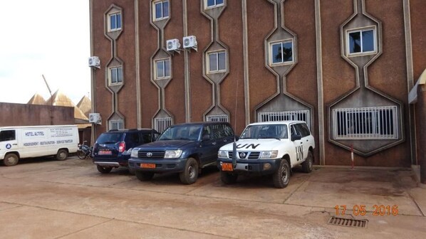 Un edificio con una fachada geométrica distintiva, que cuenta con múltiples ventanas y unidades de aire acondicionado. Hay tres vehículos estacionados al frente, incluyendo una camioneta blanca y dos SUVs.