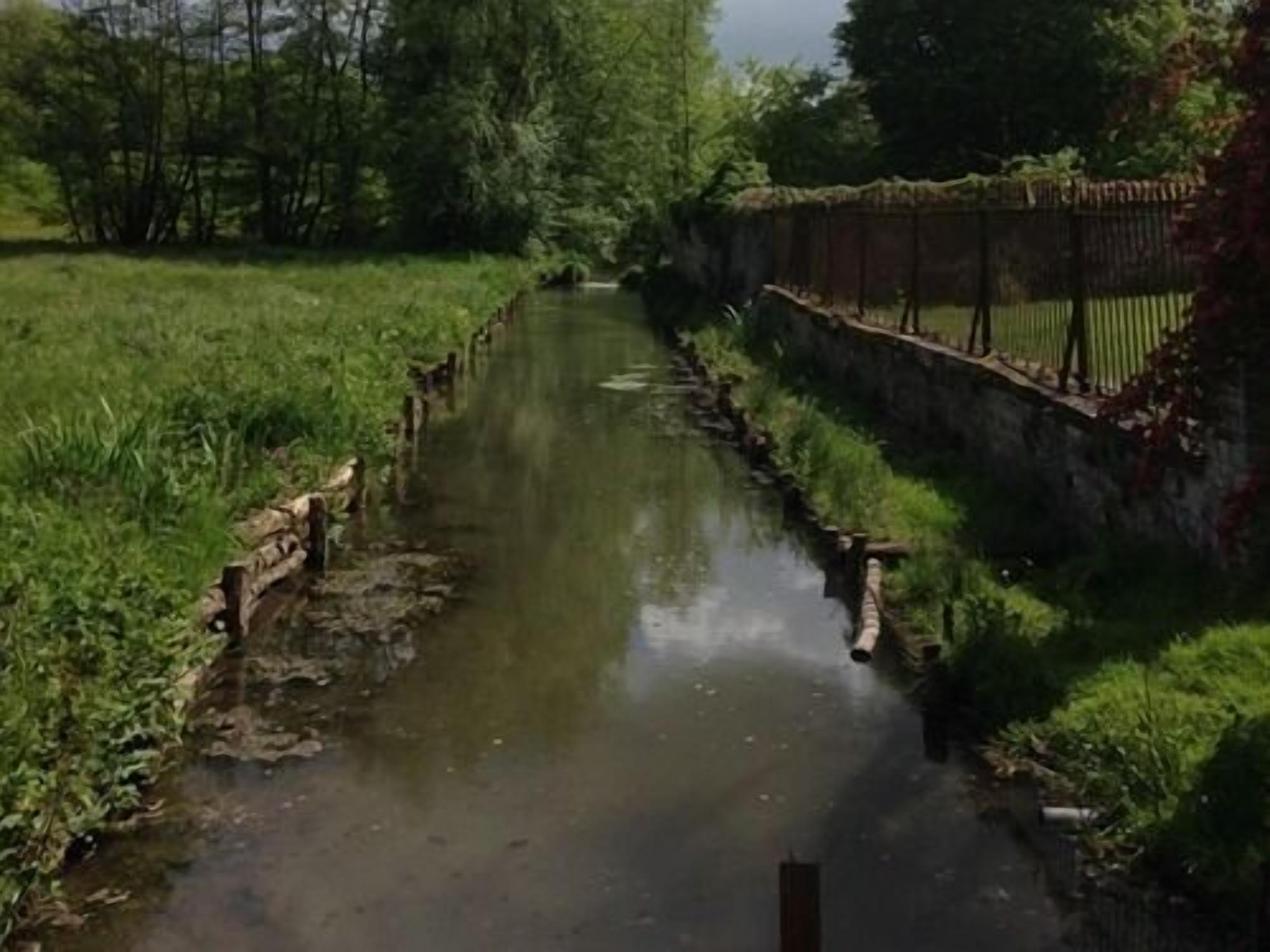 Un étroit canal bordé d’un côté par un mur de briques et de l’autre par une clôture en bois, le tout entouré de verdure.
