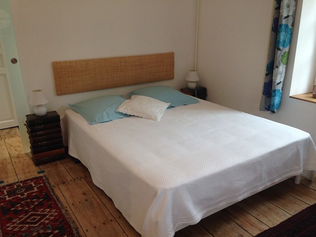 Un lit avec un drap blanc, un oreiller bleu et une tête de lit en bois. Il y a une table de chevet avec une lampe et une petite table avec une autre lampe. Un tapis à motifs est posé sur le sol.