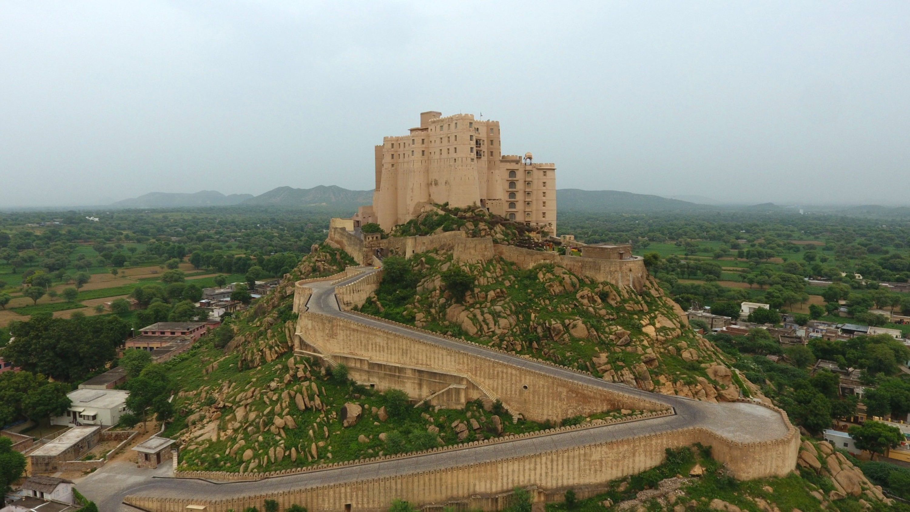 A large, historic building with a winding road leading to it, surrounded by green fields and a few scattered buildings.