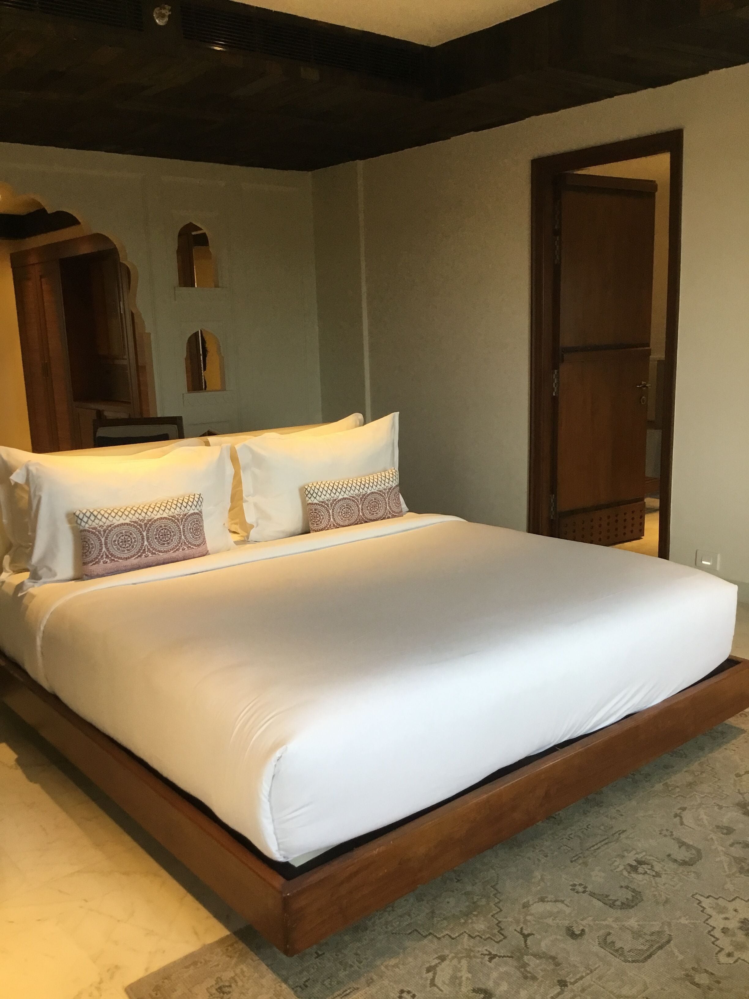 A large bed with white linens and patterned pillows, a wooden headboard, and a door to the right.