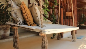 Un banco de madera rústico con cuerdas atadas a sus patas, situado en un espacio al aire libre iluminado por el sol, con una valla de madera y plantas en macetas al fondo.