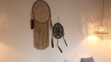 A room with two beds, each with white bedding and patterned pillows. There are woven dreamcatchers on the wall, a basket of towels, and a hanging light fixture.