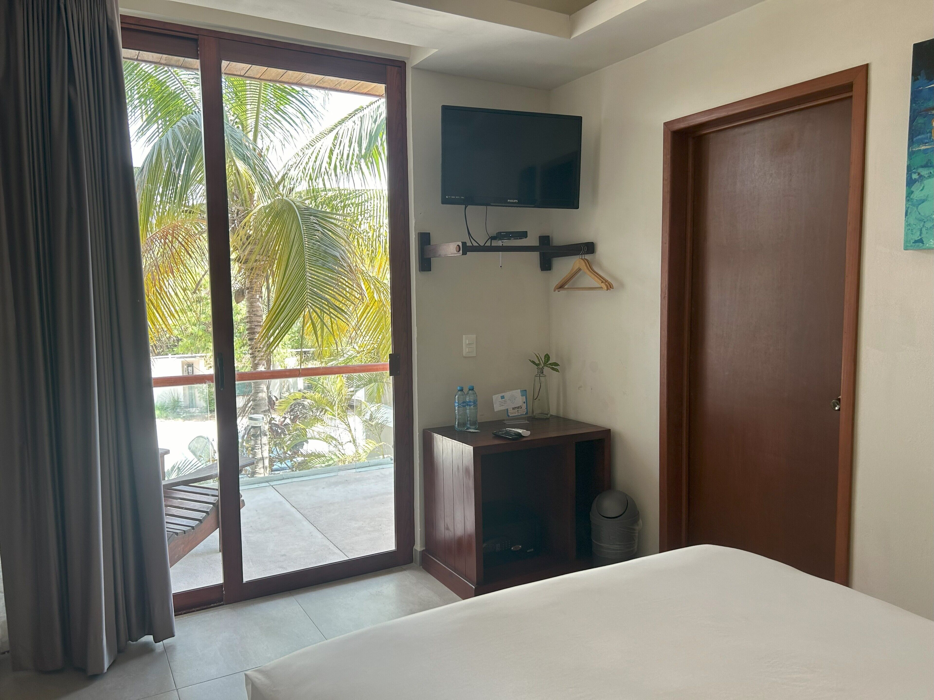 Ein Hotelzimmer mit Bett, einem an der Wand befestigten Fernseher, einem kleinen Tisch mit Flasche und Glas sowie Blick auf Palmen durch die Schiebetür.