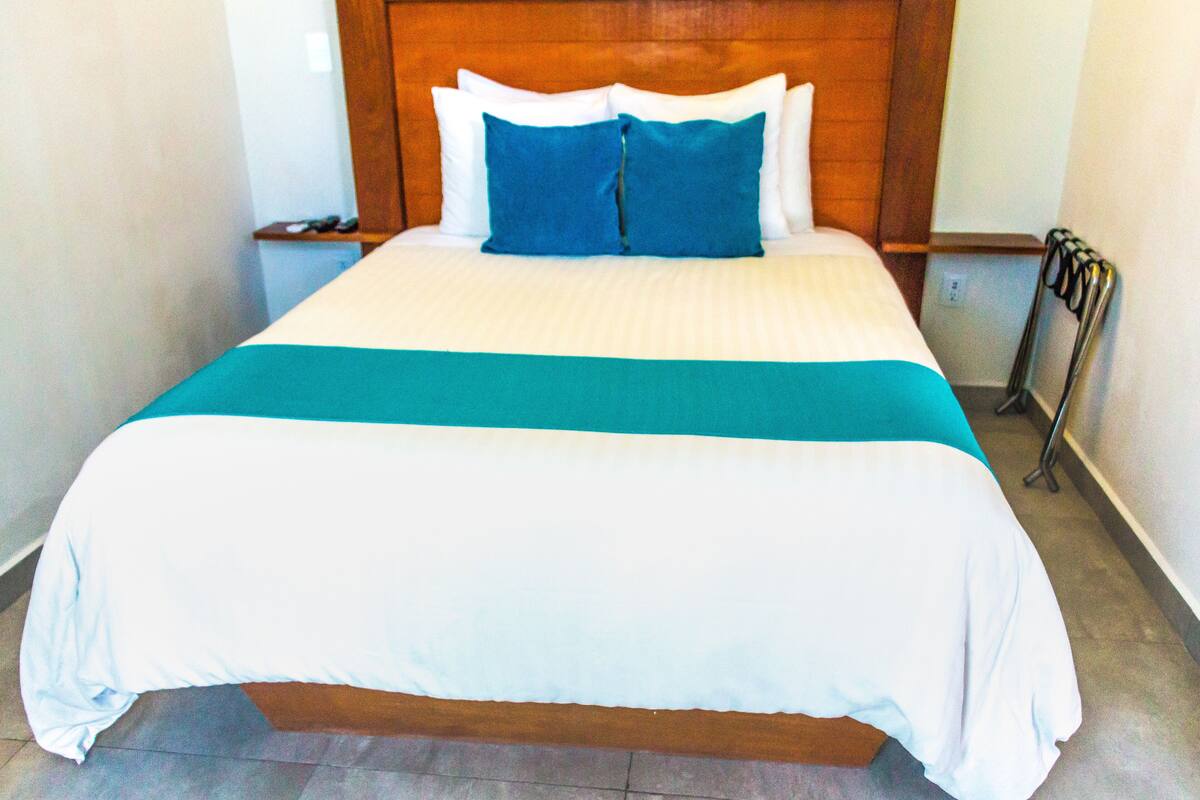 Een net opgemaakt bed met een wit- en blauwgroene sprei, een houten hoofdbord en een nachtkastje met een lamp.