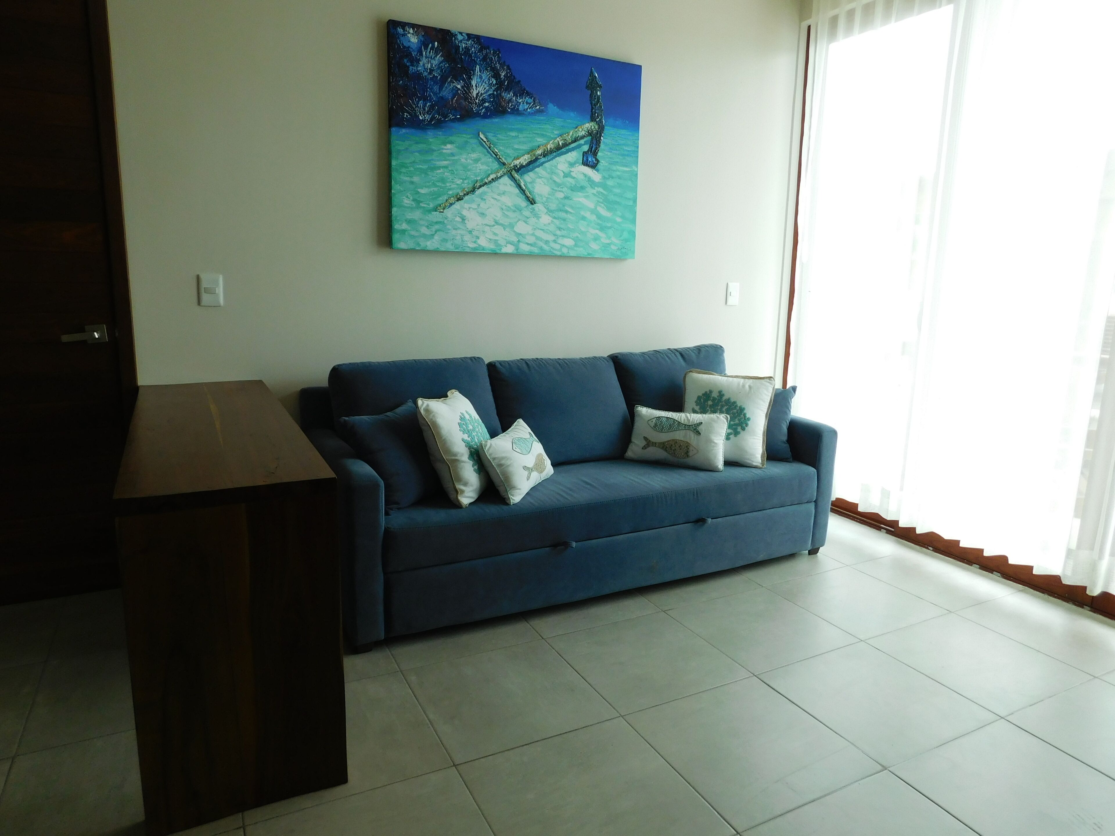 Ein Wohnzimmer mit einem dunkelblauen Sofa, zwei Zierkissen, einem Holztisch und einem Wandbild mit einer tropischen Szene.