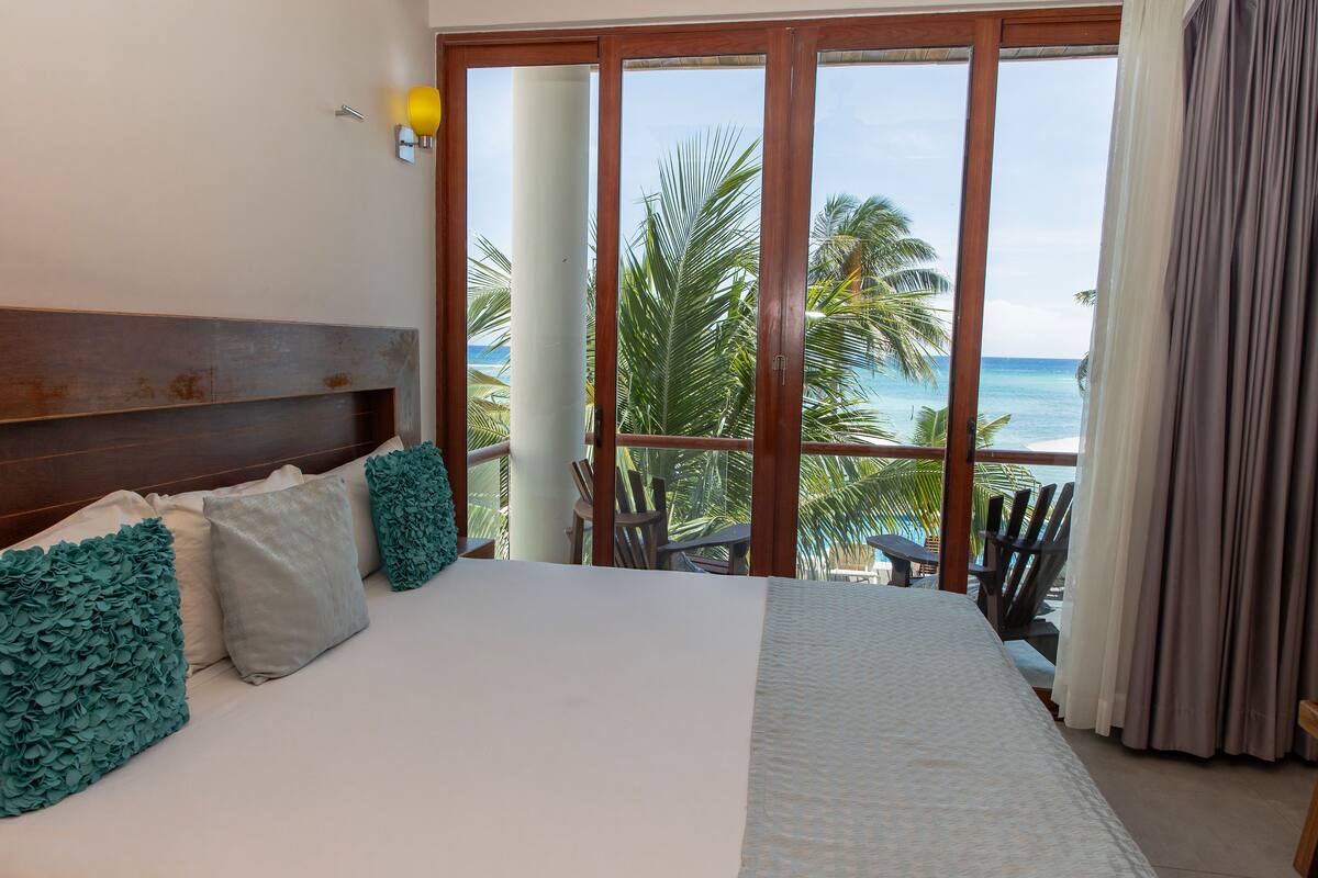 Een hotelkamer met een groot bed, een houten hoofdbord en uitzicht op de oceaan door een glazen schuifdeur.
