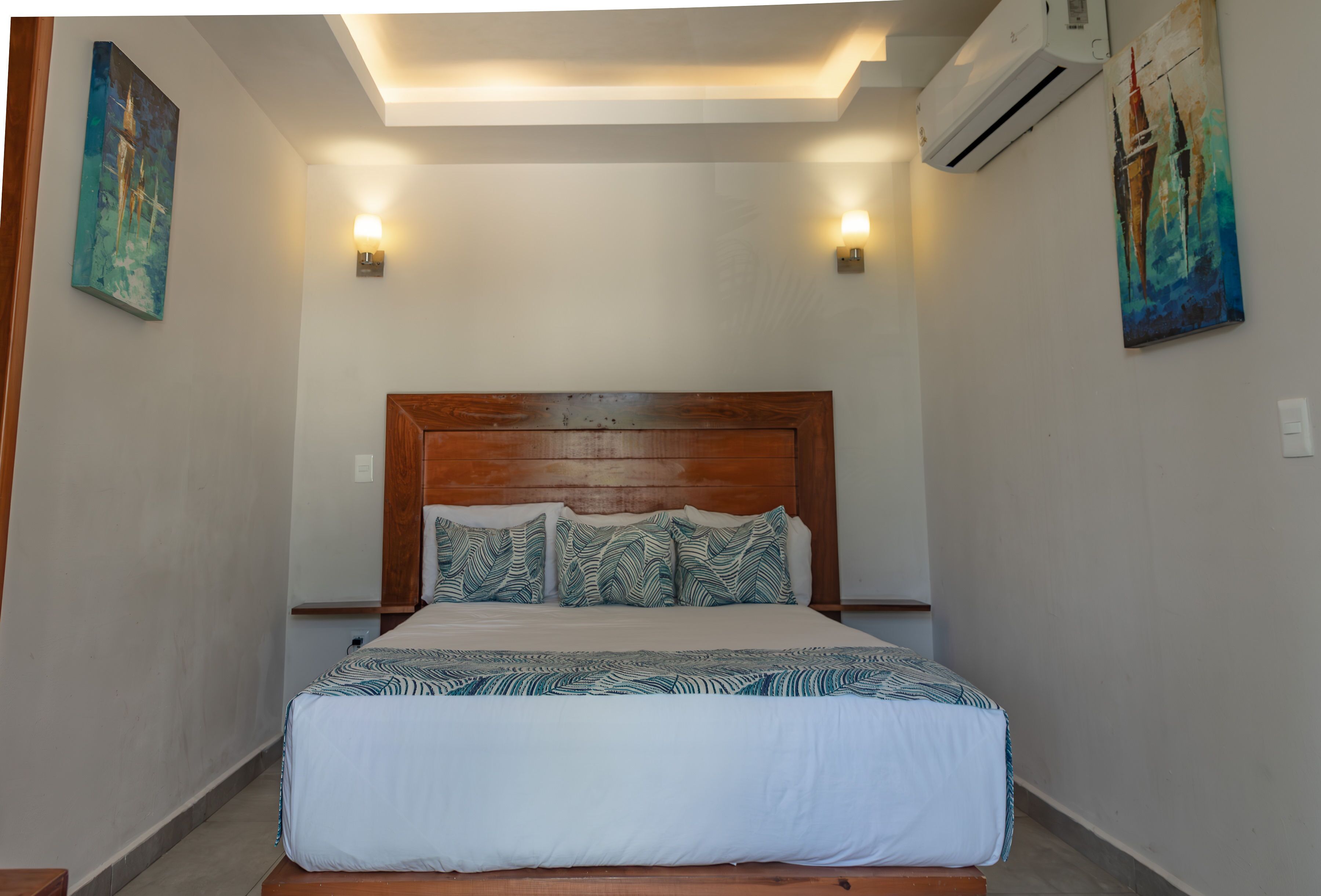 A neatly made bed with a wooden headboard, two wall-mounted lights, and two paintings on the walls.