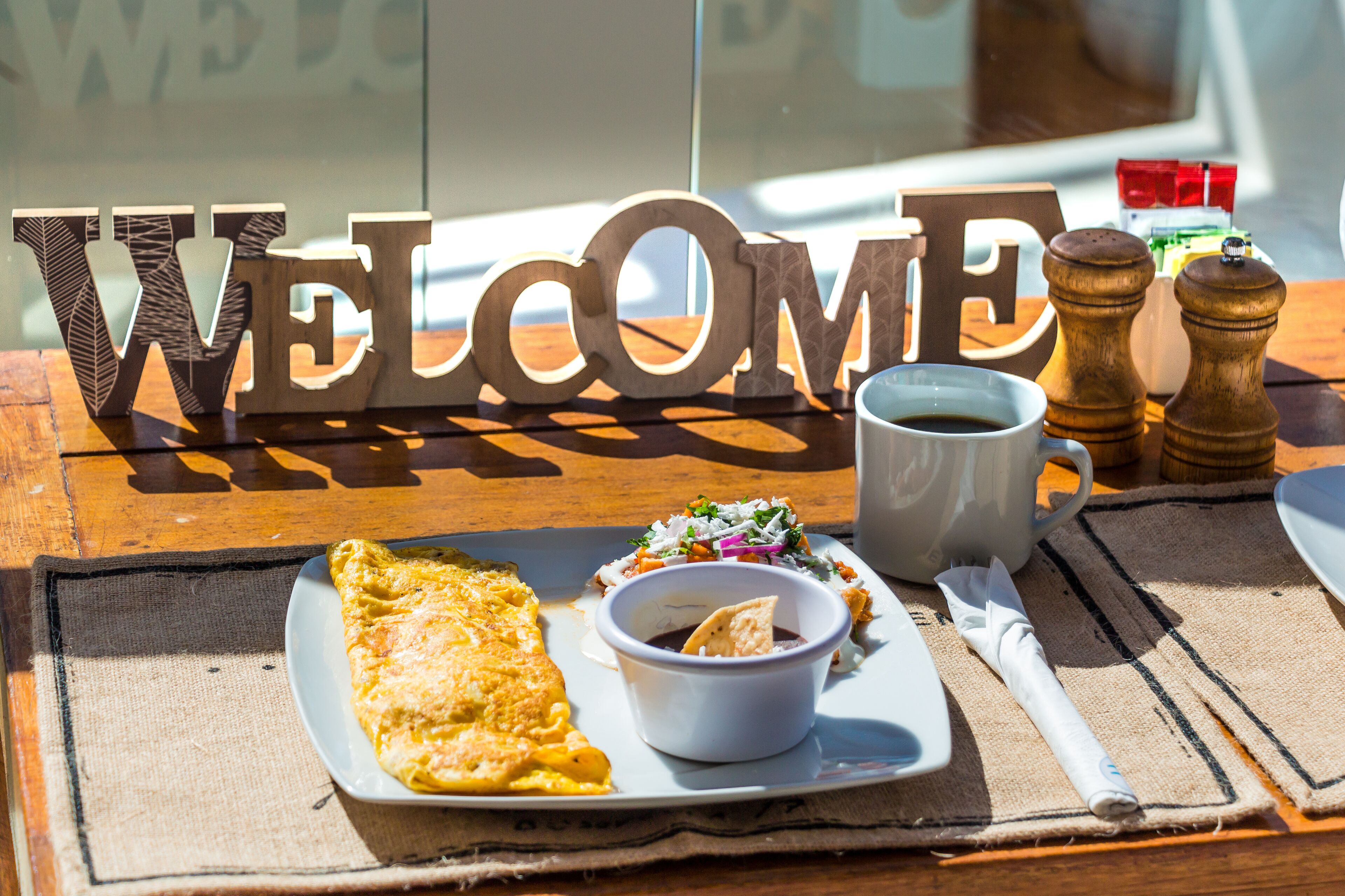 Un cartello di benvenuto con lettere di legno, un piatto con un'omelette e una ciotola di salsa, una tazza di caffè e dei condimenti da tavola.