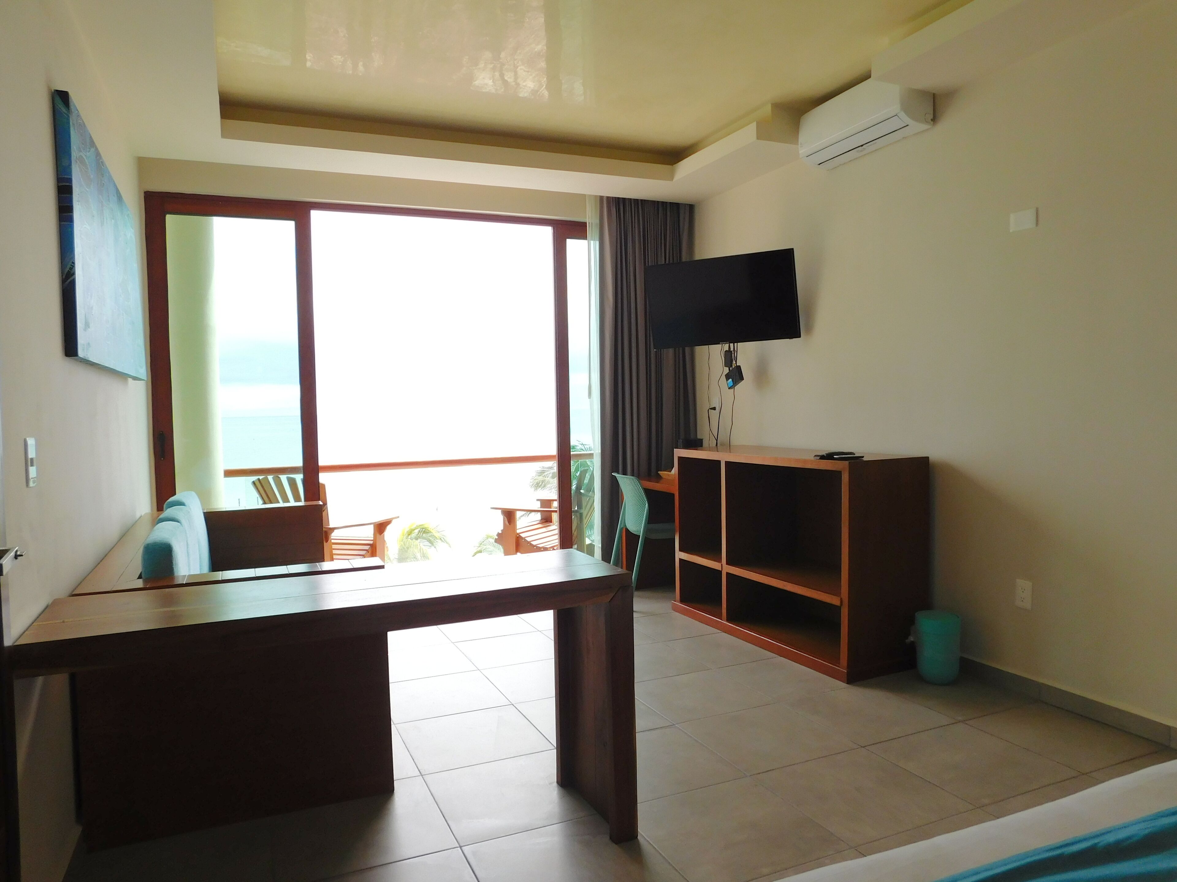 Habitación con una ventana grande, un televisor en la pared, un escritorio de madera y un pequeño rincón de estar con sillas.
