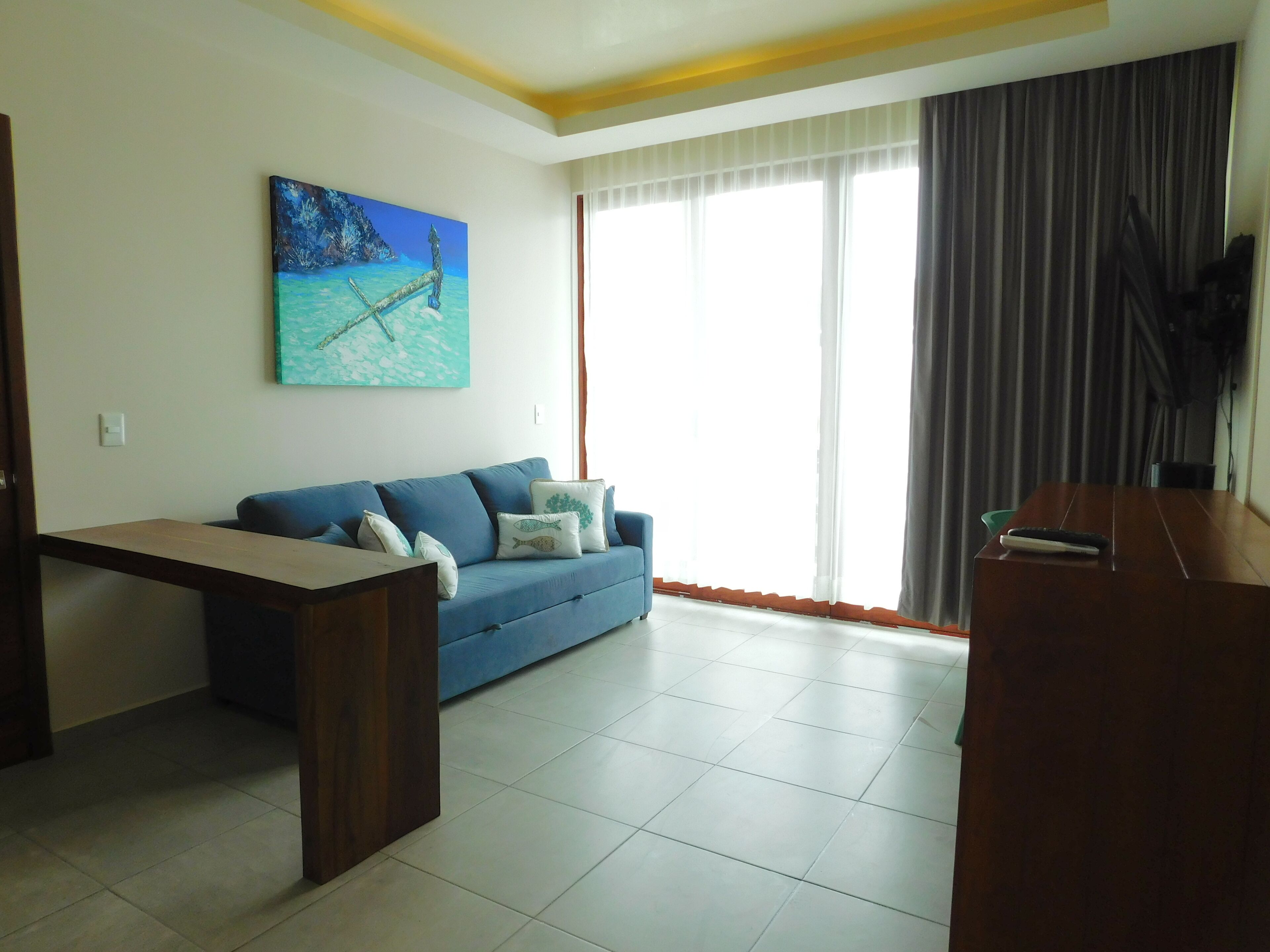 A living room with a blue sofa, a painting of a seascape, and a large window with curtains.