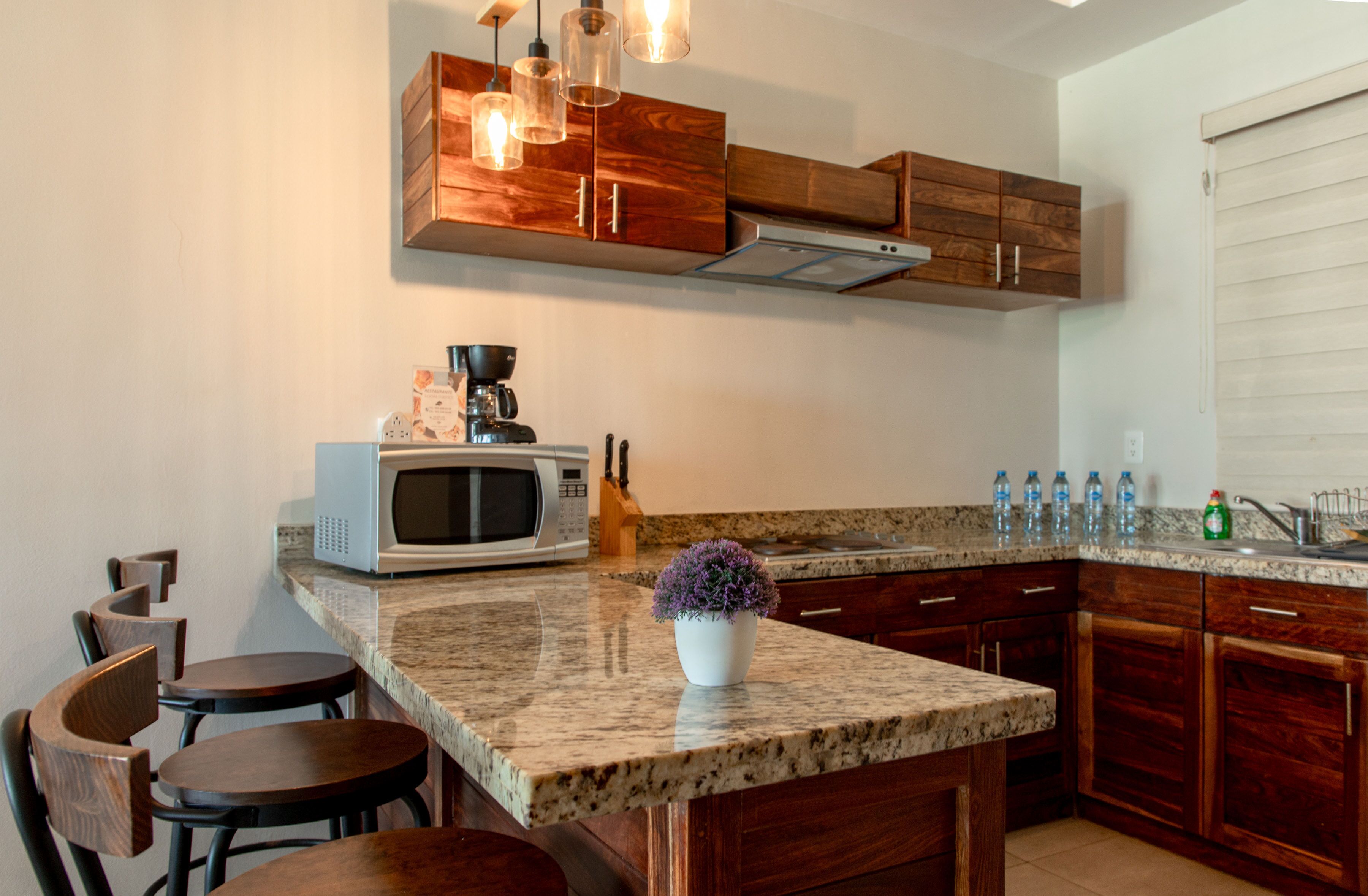 A kitchen with granite countertops, wooden cabinets, a built-in microwave, and a coffee maker. There are bar stools and a potted plant on the counter.