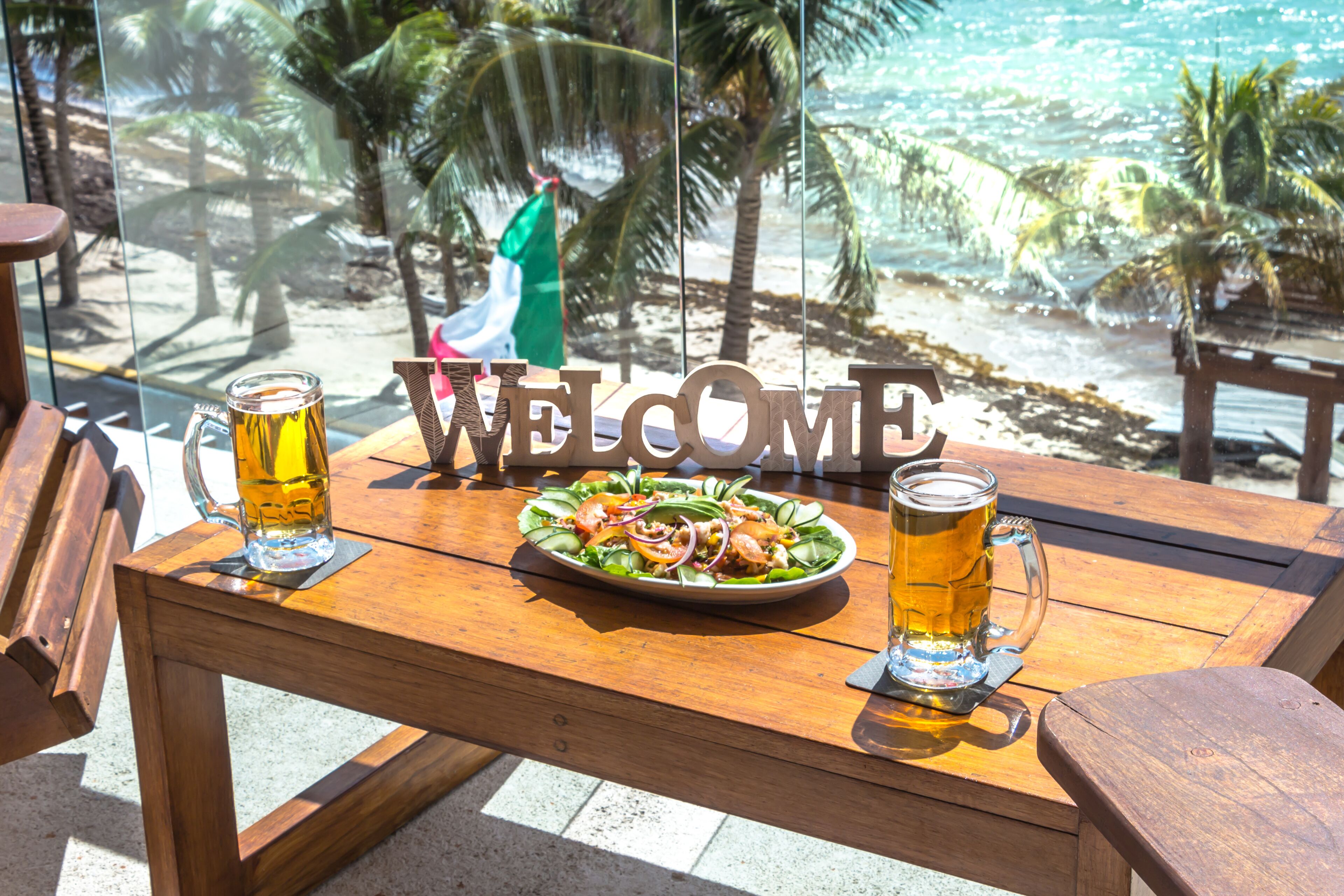 A wooden table with a welcome sign, two beer mugs, and a plate of food.