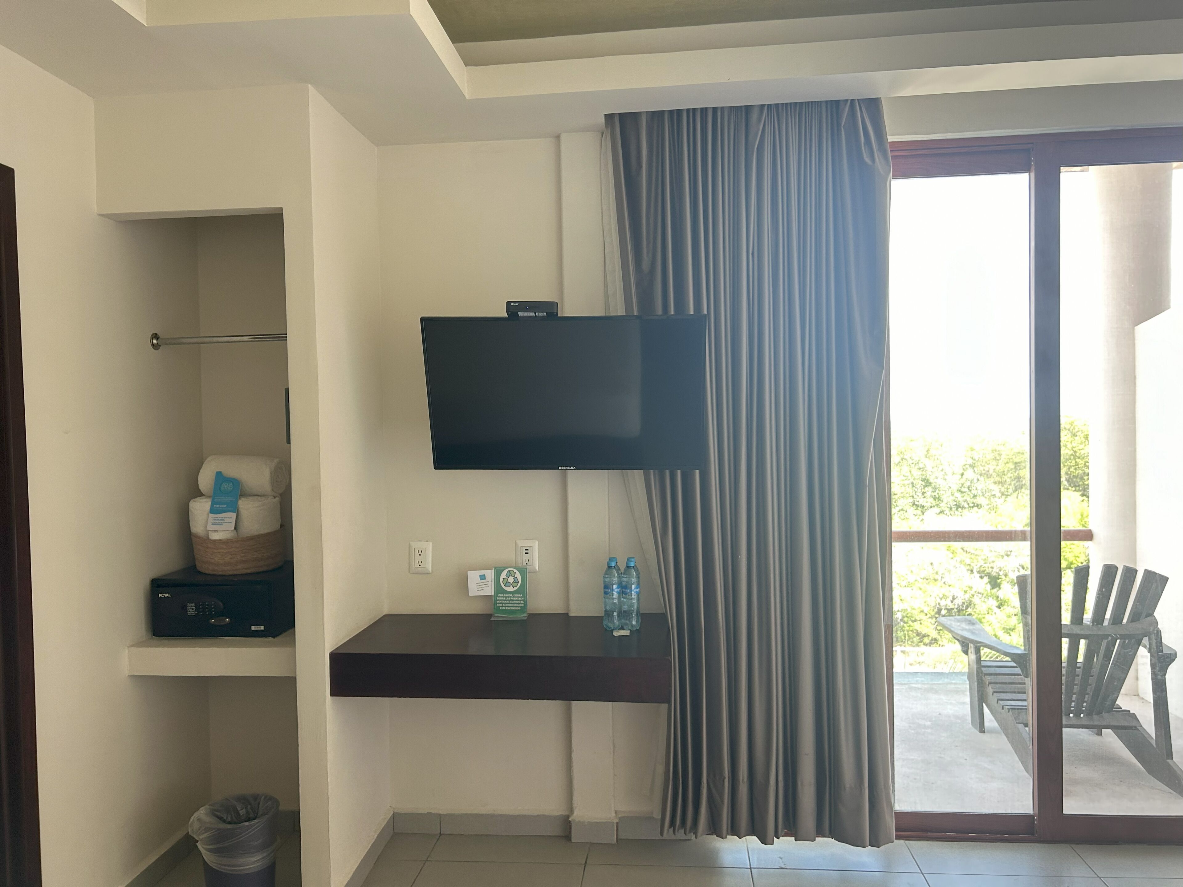 Habitación de hotel con televisión de pantalla plana en la pared, un estante pequeño con artículos de aseo, un cesto de basura y vistas del patio exterior a través de una puerta corrediza de vidrio.
