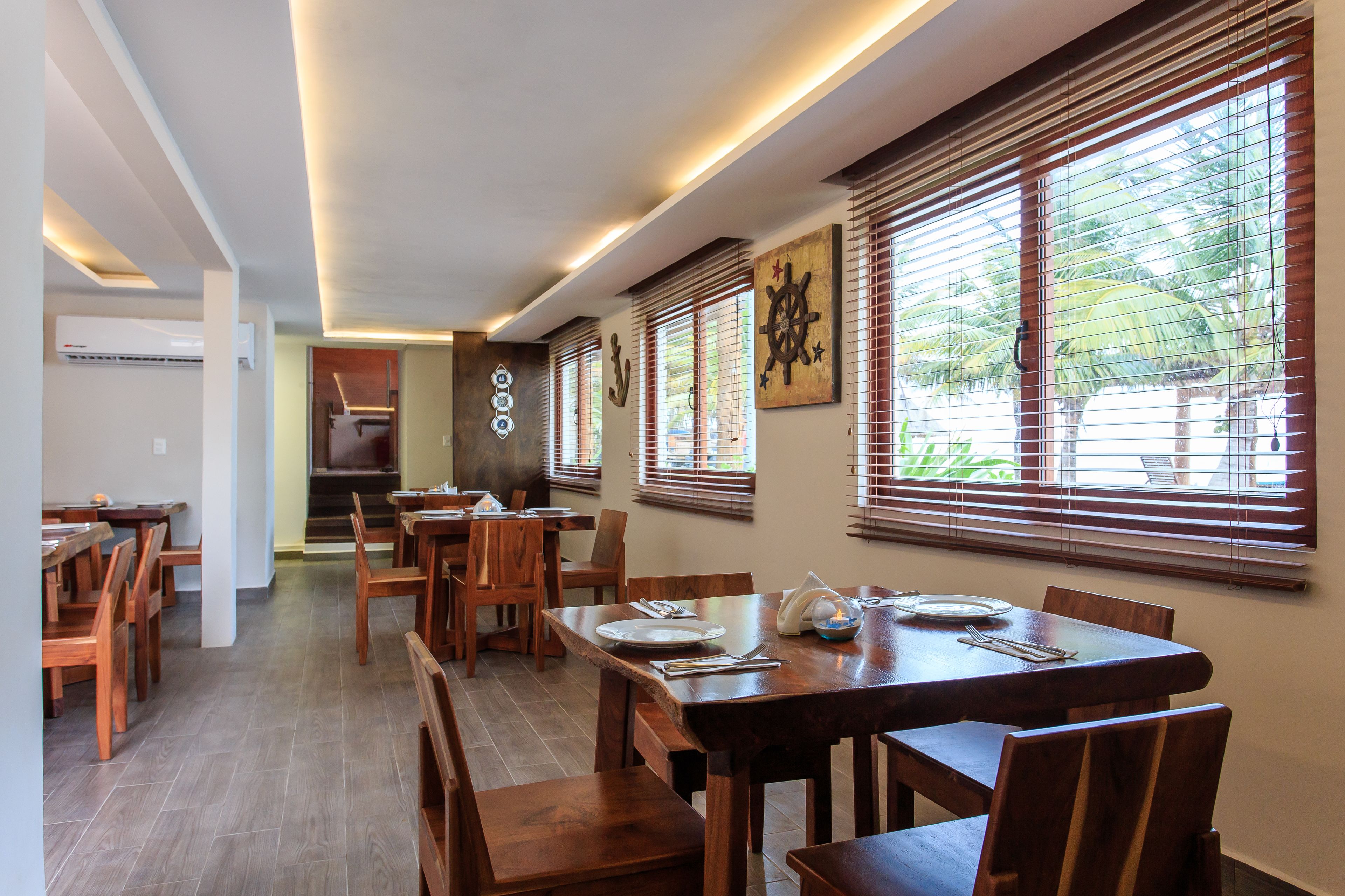 Un espace repas avec des tables et des chaises en bois, de grandes fenêtres équipées de stores et des œuvres d’art murales, dont une représentation d’un gouvernail de navire.