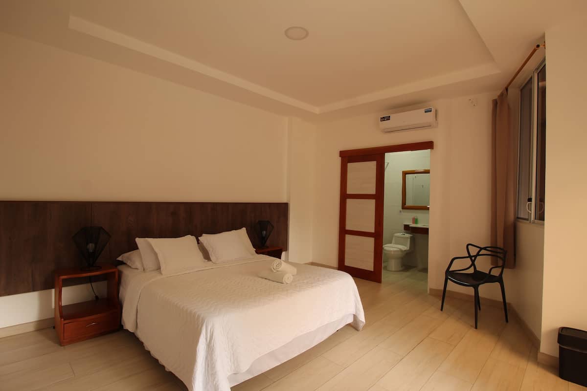 Un dormitorio con cabecera de madera, cama con ropa blanca, mesita de noche, una silla y un equipo de aire acondicionado.