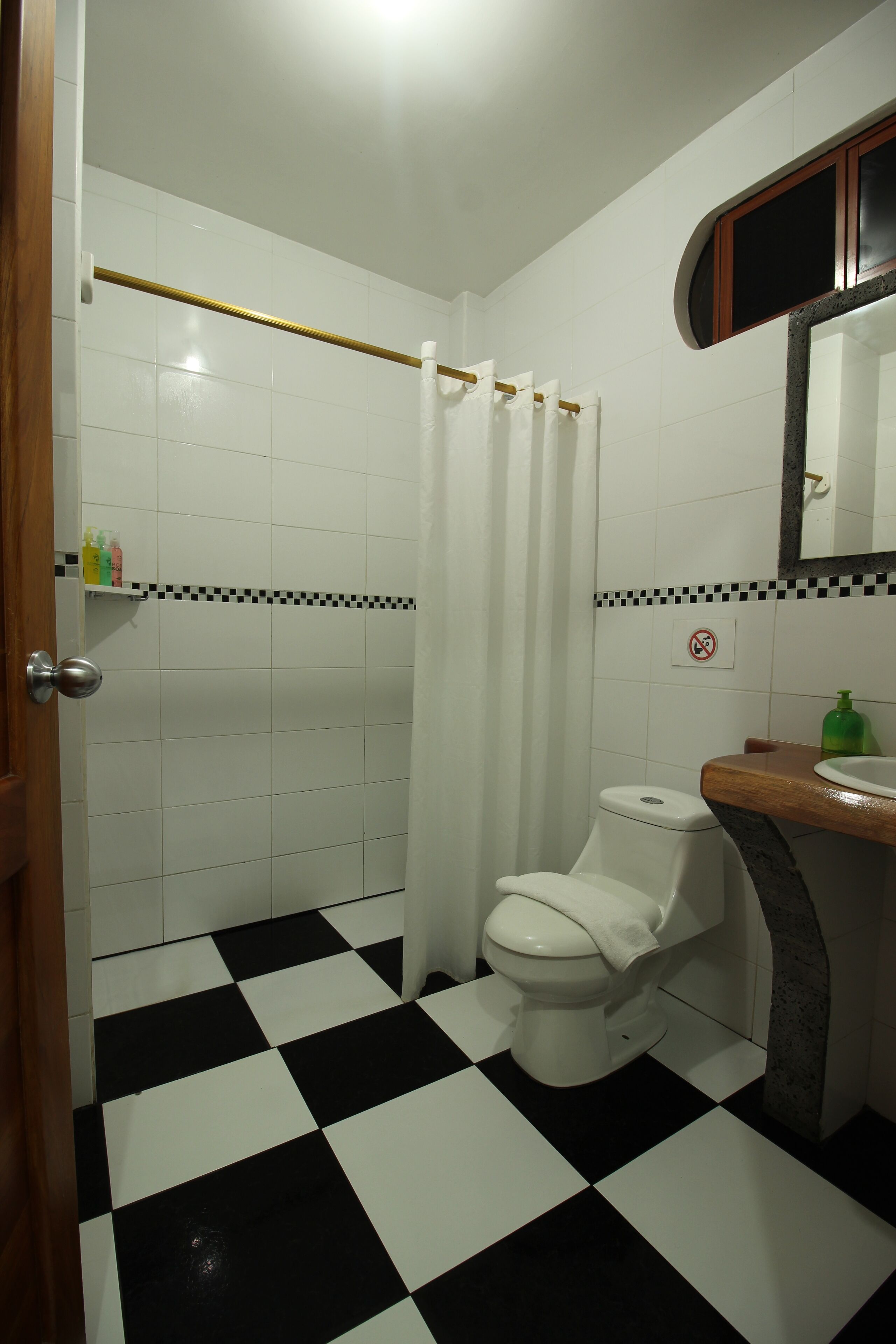Een badkamer met een zwart-wit geruite vloer, een wit toilet, een douchegordijn en een houten wastafelmeubel met spiegel.