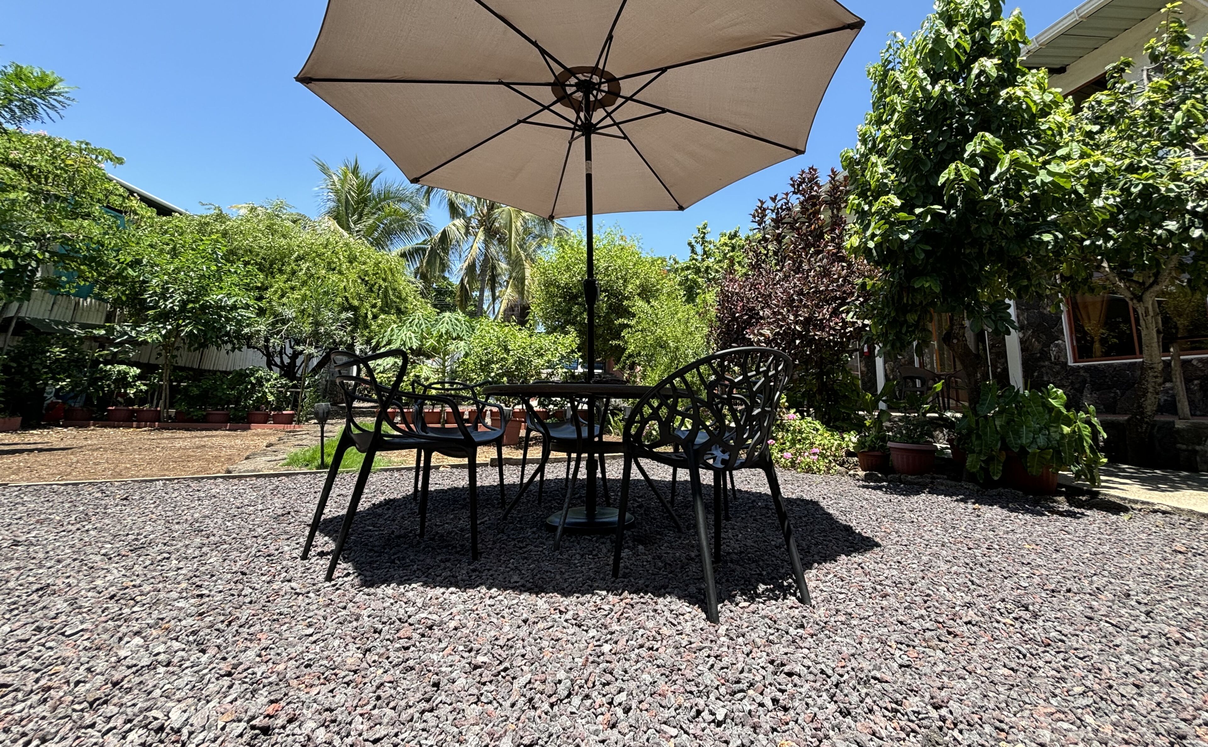 Un espacio al aire libre con mesa y sillas bajo un gran paraguas, rodeado de grava y plantas en macetas.