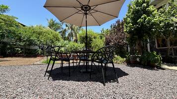 Un espacio al aire libre con mesa y sillas bajo un gran paraguas, rodeado de grava y plantas en macetas.