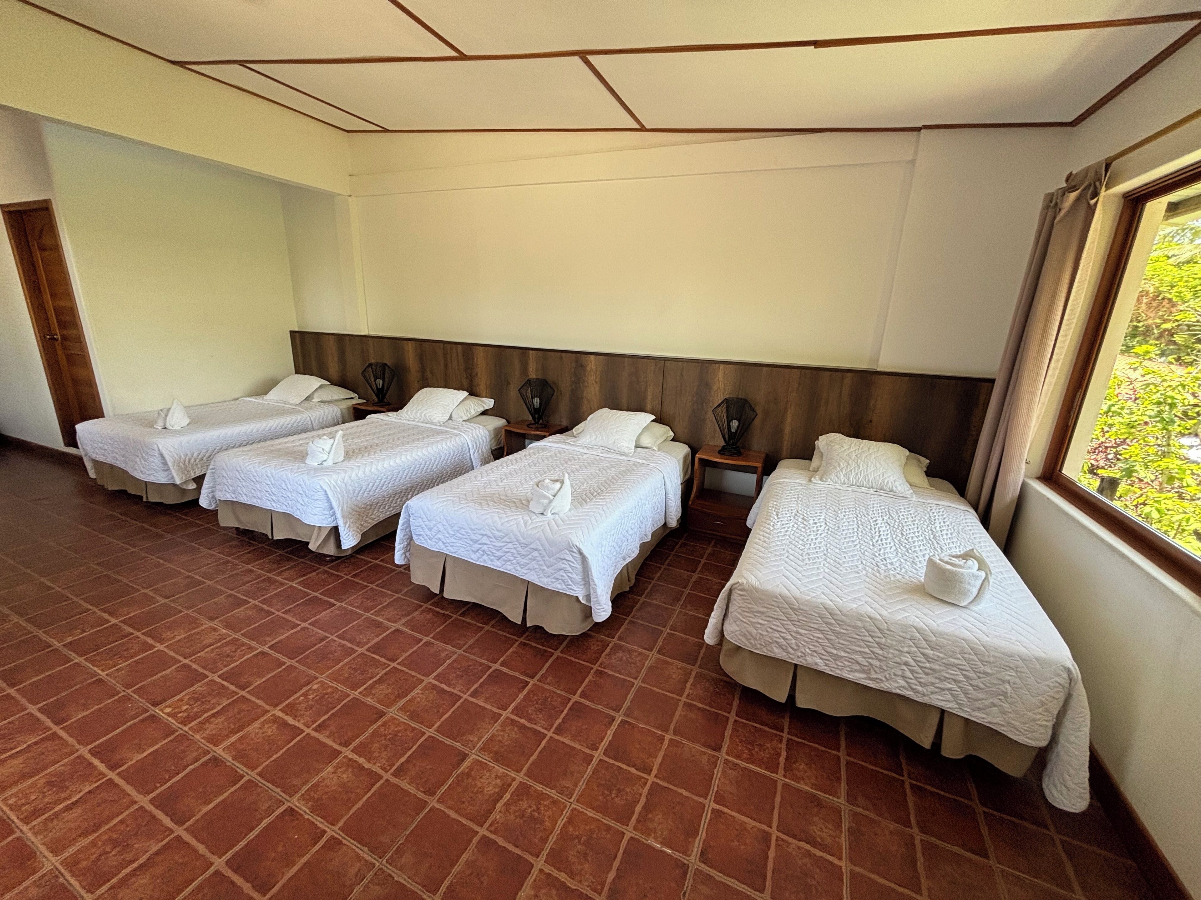 A hotel room with four single beds, a wooden headboard, a tiled floor, and a window with a view of greenery.