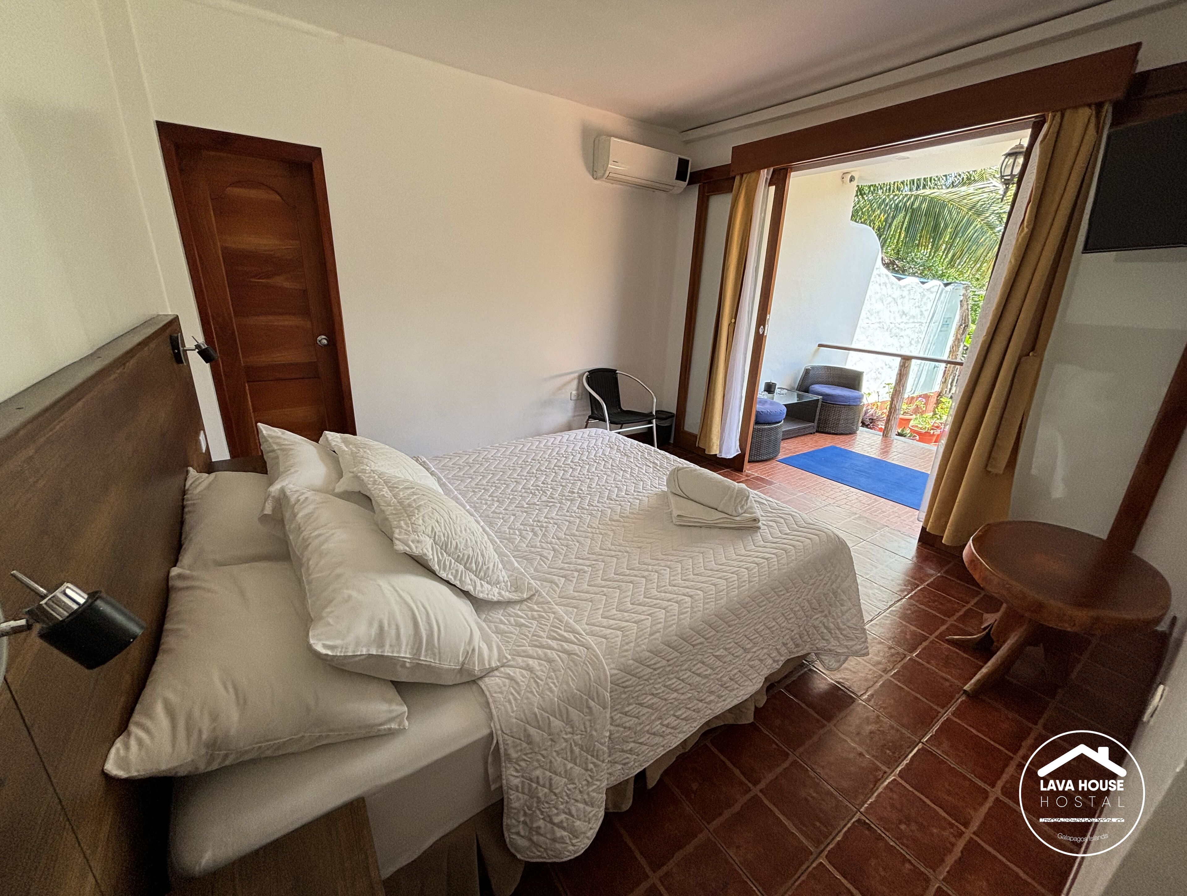 A bedroom with a bed, pillows, a wooden headboard, a small table, a chair, and a view of the outside.
