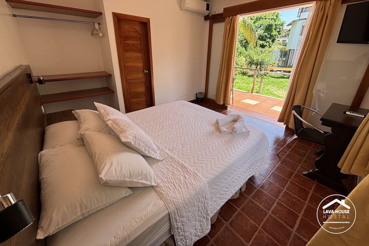 Habitación de hotel con cama, cabecera de madera, escritorio pequeño con silla, televisor y ventana con cortinas.