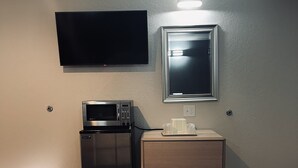 A hotel room with a flat-screen TV mounted on the wall, a microwave, a refrigerator, a wooden dresser, and a bedside table with a cup and a trash bin. - Century 21 Motel (Las Cruces)