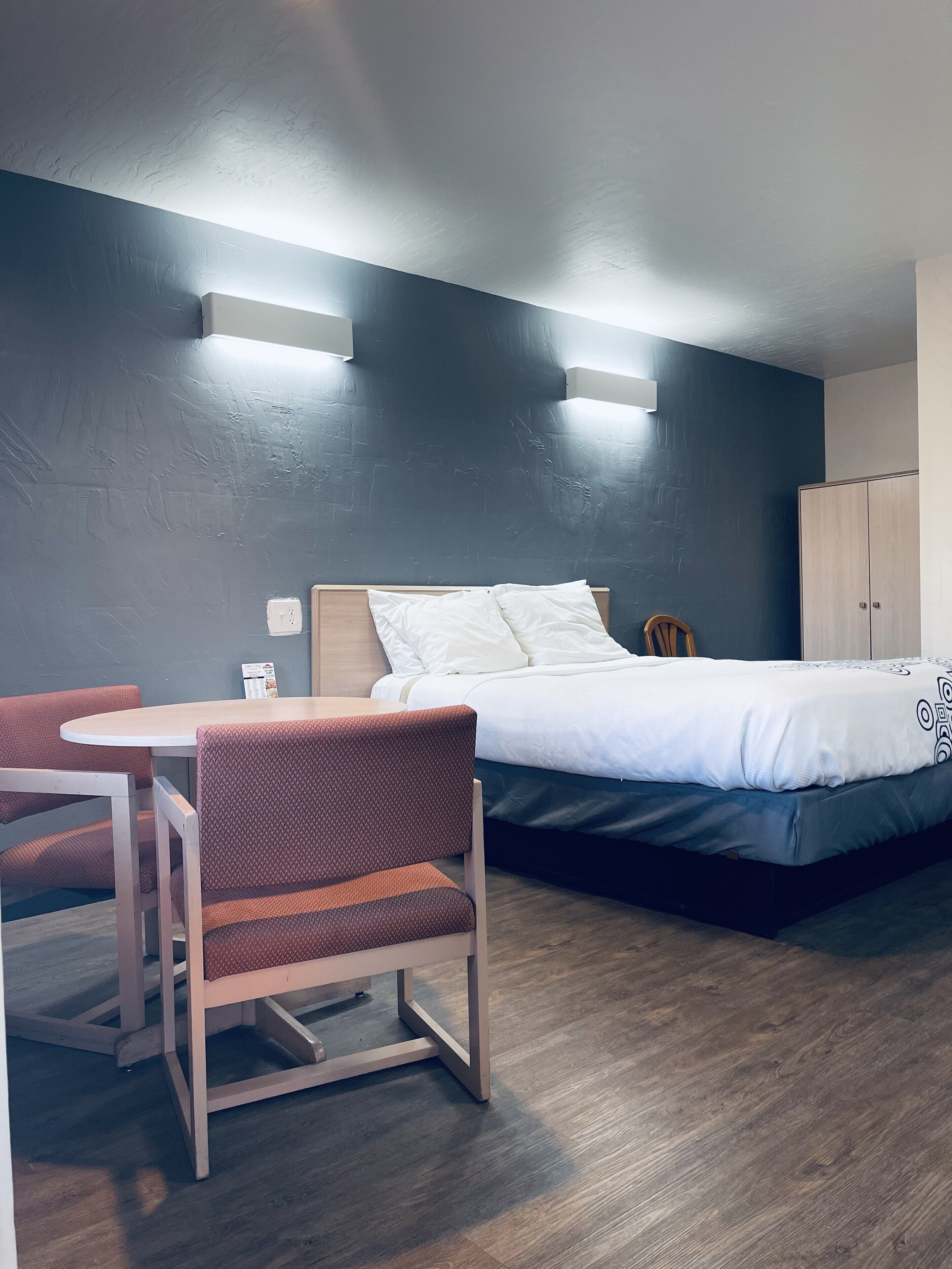 Habitación de hotel con cama, una mesita pequeña con dos sillas, un armario de madera y una lámpara fijada en la pared.