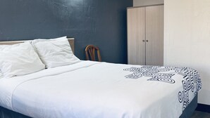 A neatly made bed with white linens and a geometric-patterned blanket, a wooden chair, and a wardrobe in a room with dark walls and a light-colored floor. - Century 21 Motel (Las Cruces)