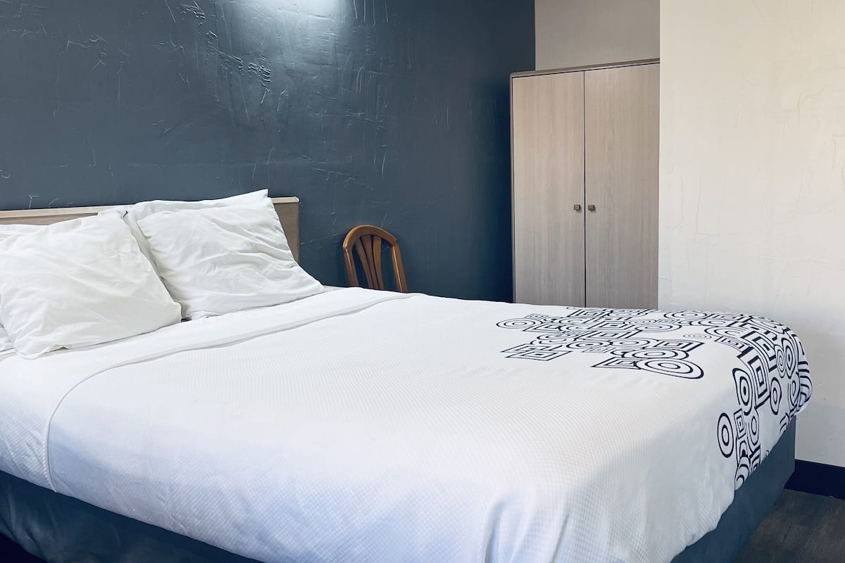 A neatly made bed with white linens and a geometric-patterned blanket, a wooden chair, and a wardrobe in a room with dark walls and a light-colored floor.