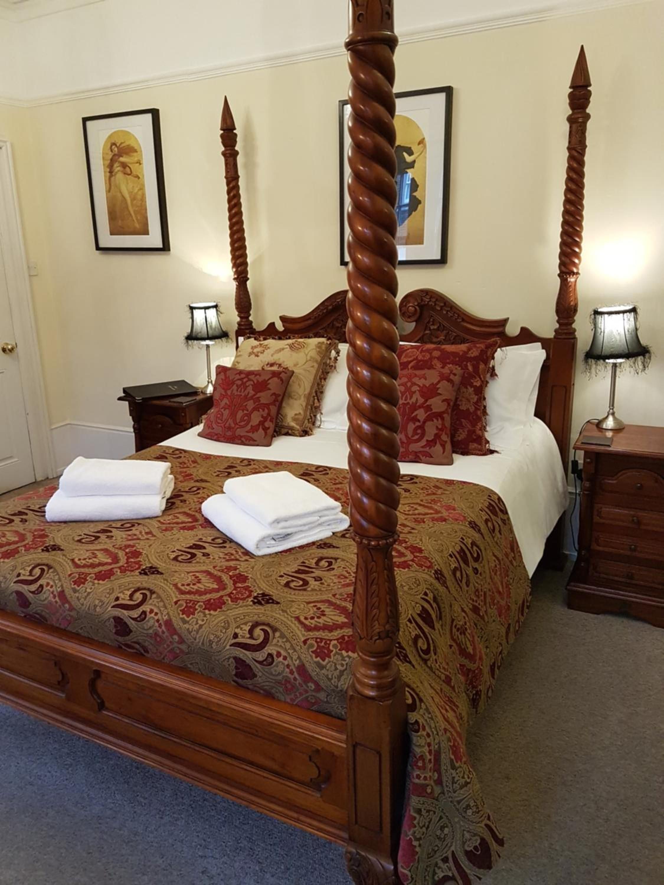 A four-poster bed with a wooden frame and a patterned bedspread. There are two bedside tables with lamps, and a door is visible to the left.
