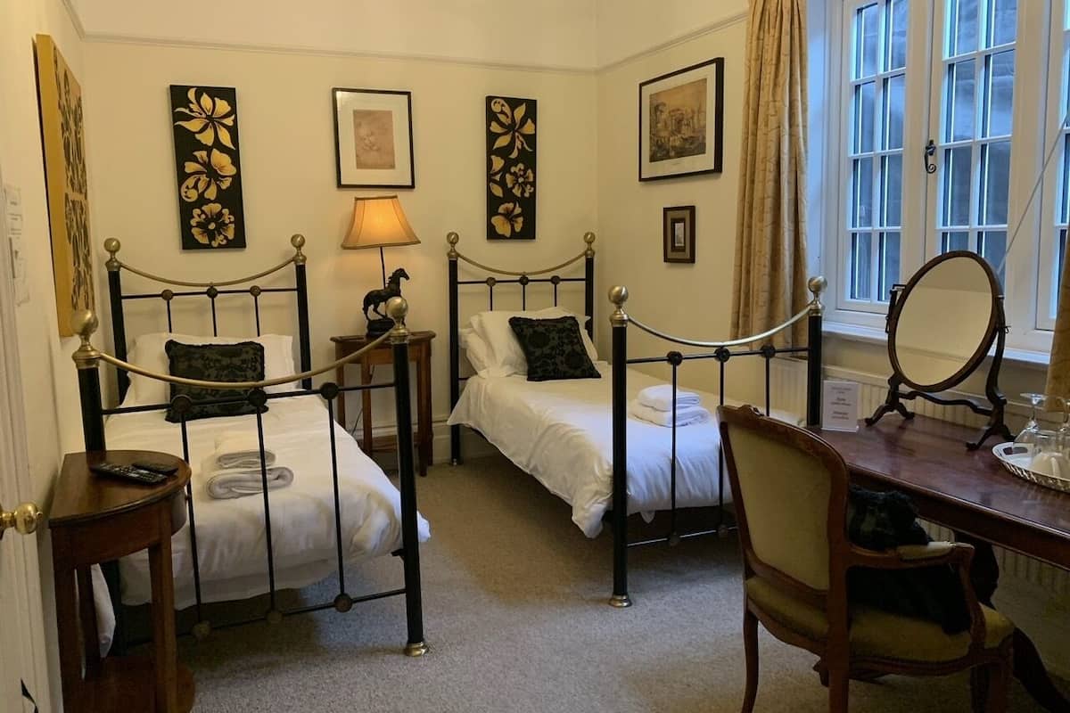 A hotel room with two single beds, a wooden desk, a chair, a lamp, and artwork on the walls.
