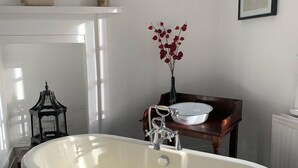 Un baño con una bañera independiente, una mesita con un cuenco y un jarrón de flores rojas, y un cuadro en la pared.