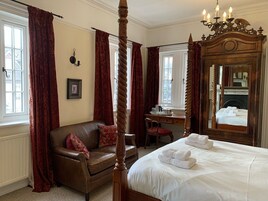 Uma cama de dossel tradicional com roupa de cama branca, um sofá de couro com almofadas vermelhas, uma escrivaninha de madeira com uma cadeira e um espelho grande.