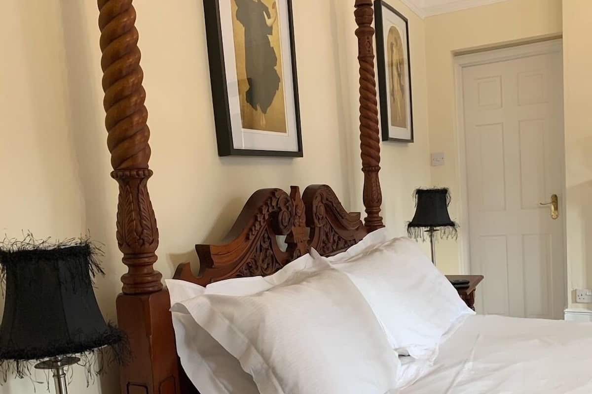 A four-poster bed with white linens, a wooden nightstand with a lamp, and two framed pictures on the wall.