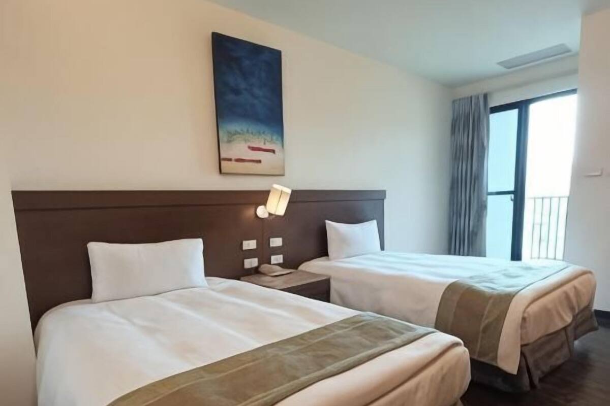 A hotel room with two beds, a nightstand, a painting on the wall, and a window with curtains.