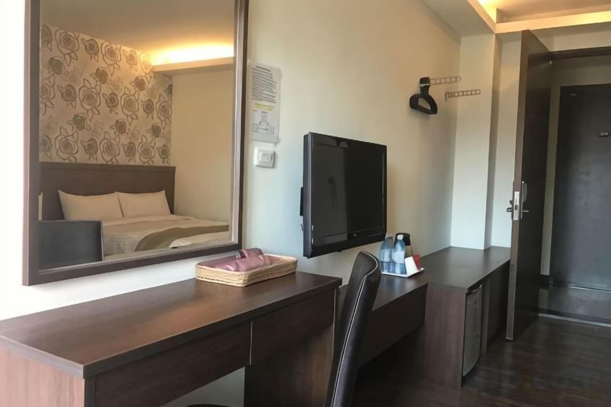 A hotel room with a large mirror, a flat-screen TV, a desk with a chair, and a bed with a headboard.