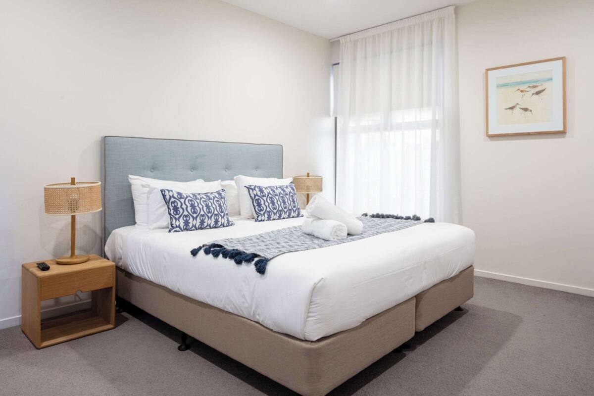 A neatly made bed with a grey headboard, white bedding, and blue patterned pillows. There is a small wooden nightstand with a lamp beside the bed.