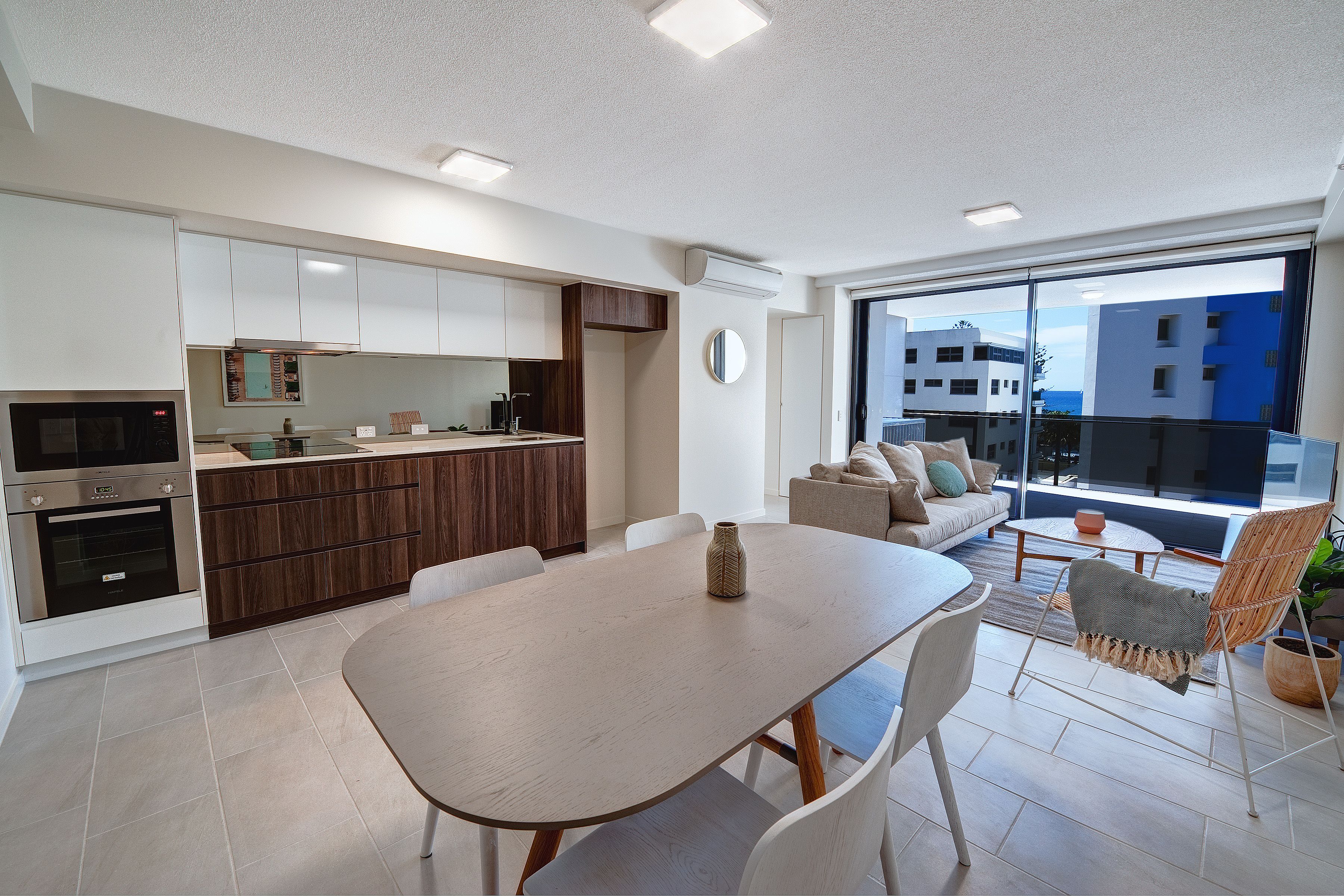 Cozinha moderna com eletrodomésticos embutidos, área de jantar com mesa e cadeiras, e varanda com vista para prédios.
