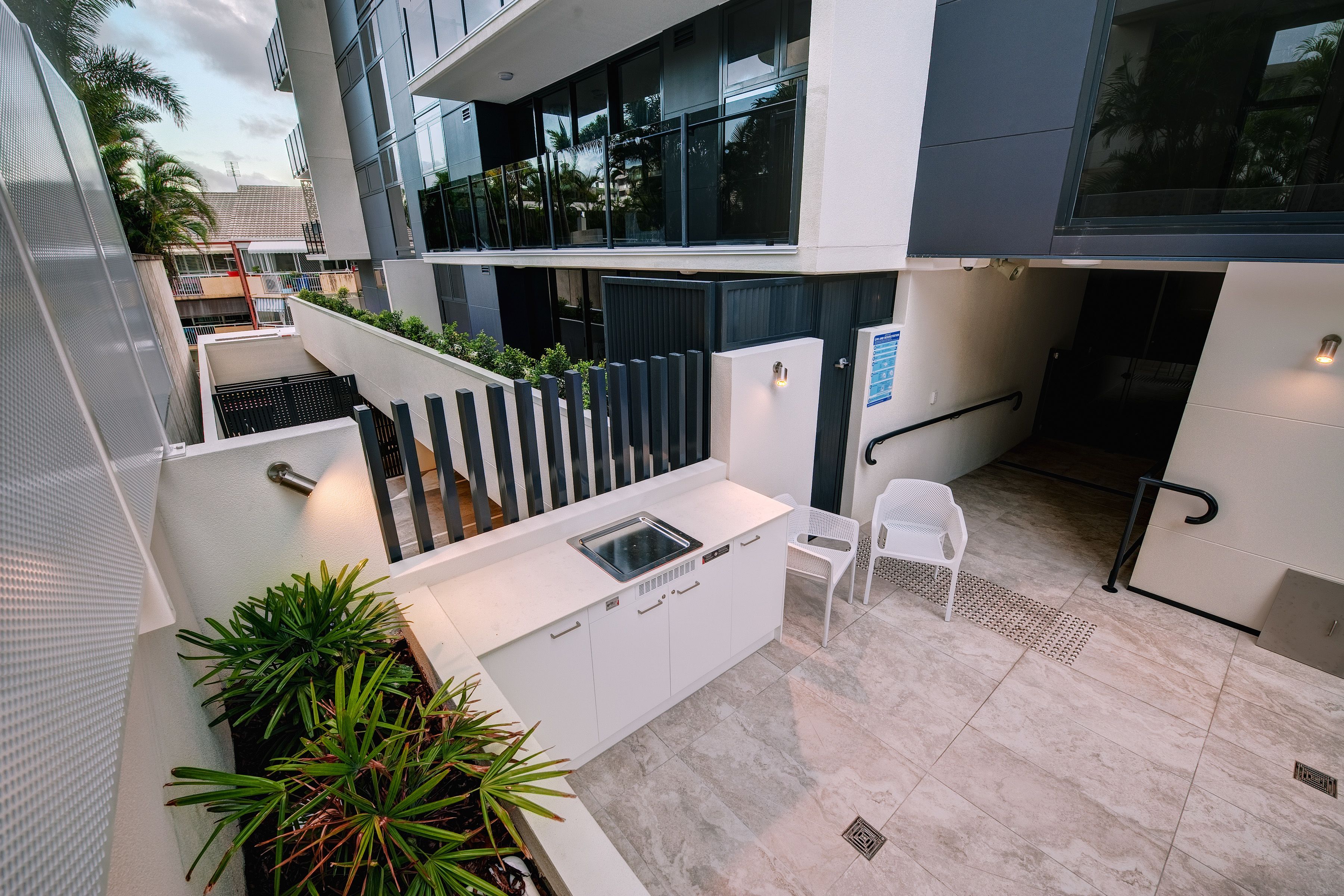 Un espacio moderno de cocina al aire libre con un lavamanos y una silla blanca, rodeado de plantas en macetas y un piso de baldosas.