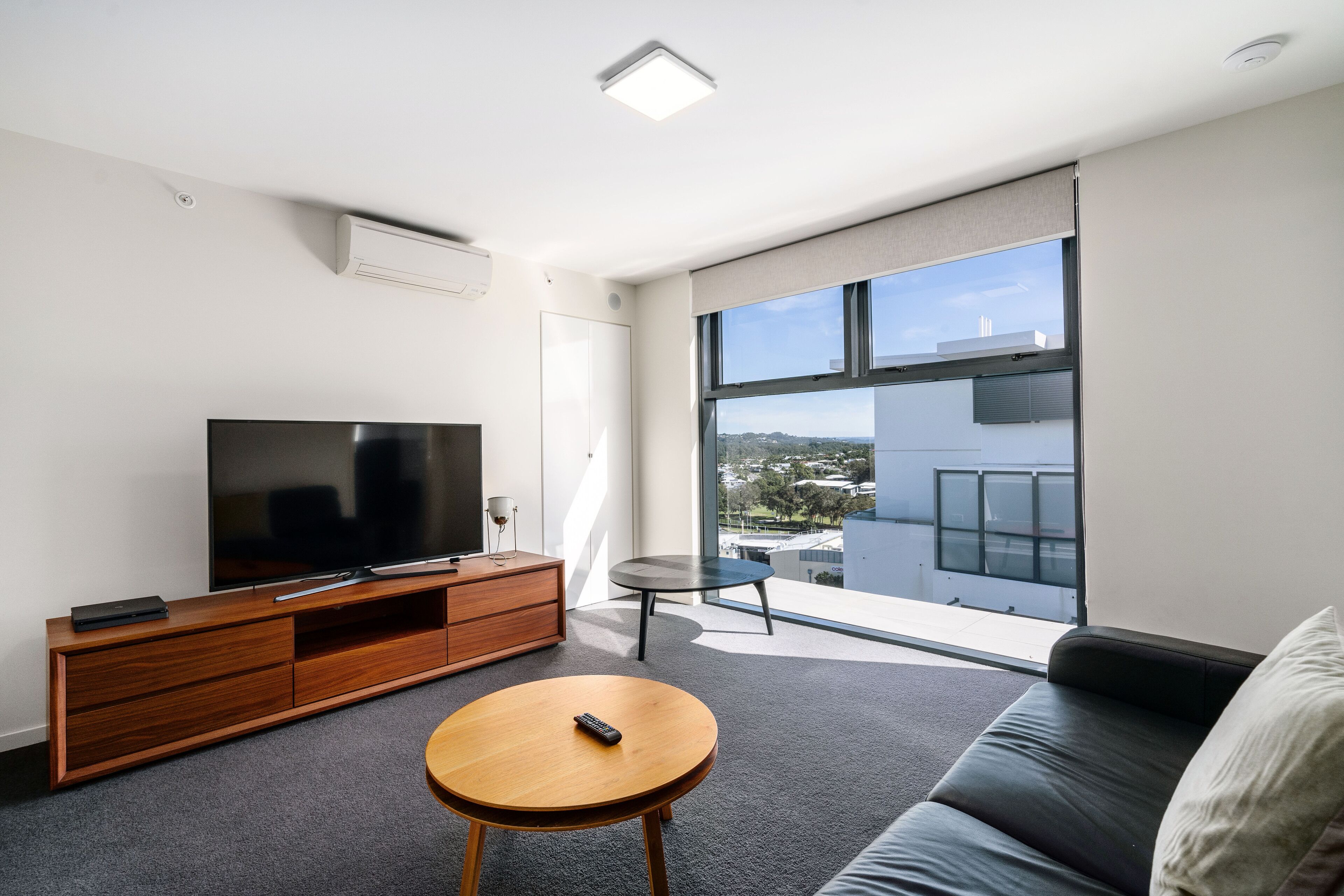 Una sala moderna con una televisión de pantalla plana, mueble de madera para entretenimiento, una mesa de centro redonda, un sofá de cuero negro y un amplio ventanal con vista a la ciudad.
