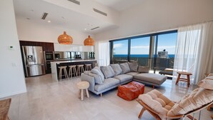 A modern living room with a grey sectional sofa, a wooden bench, and a cushioned chair. A kitchen with a bar area is visible in the background. - First Light Mooloolaba, an Ascend Collection Hotel (Mooloolaba)