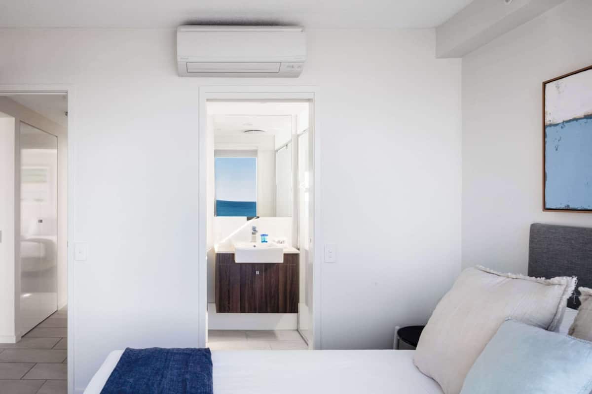 A bedroom with a bed, a bedside table, a nightstand, a wall-mounted air conditioner, a bathroom with a sink and mirror, and a view of the sea through the window.