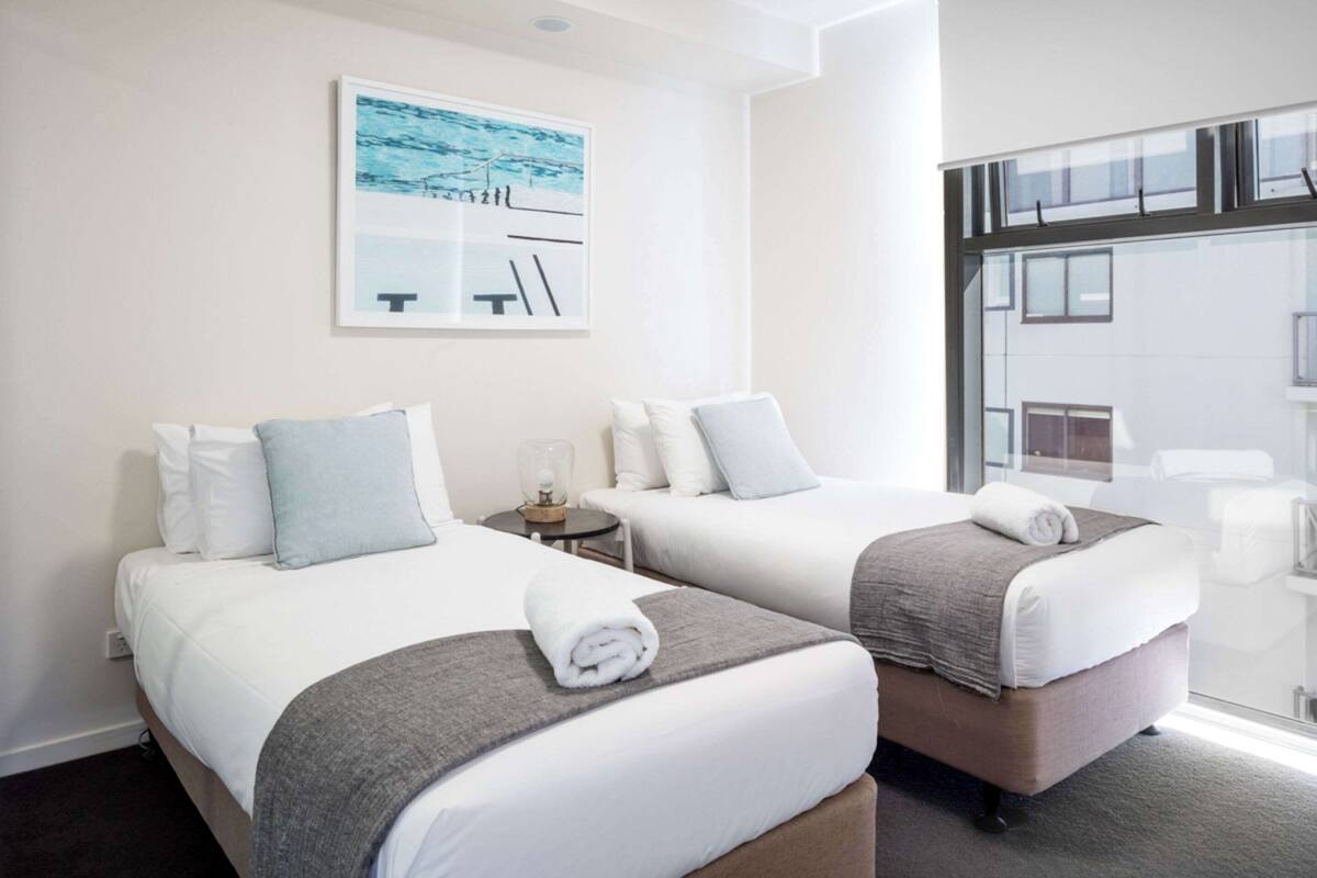A hotel room with two beds, a small table, a wall-mounted artwork, and a large window.