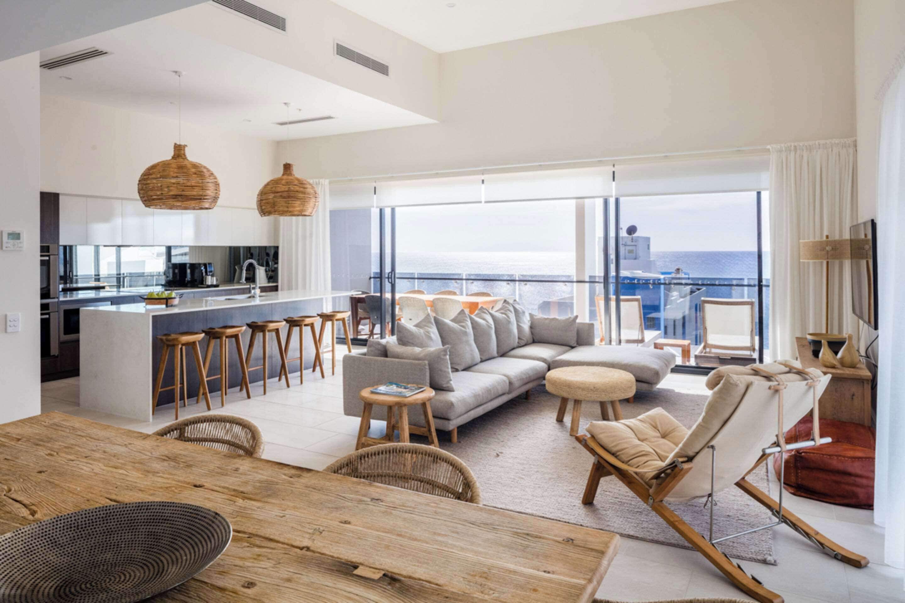 A modern living room with a sofa, chairs, and a dining area with a wooden table. There is a kitchen with a bar and large windows offering a view of the sea.