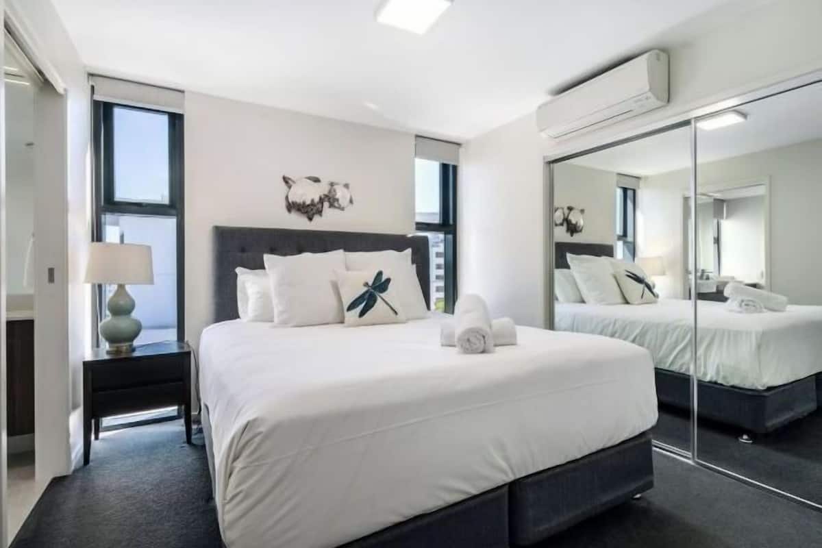 A modern hotel room with a large bed, a nightstand with a lamp, a mirror, and a window.