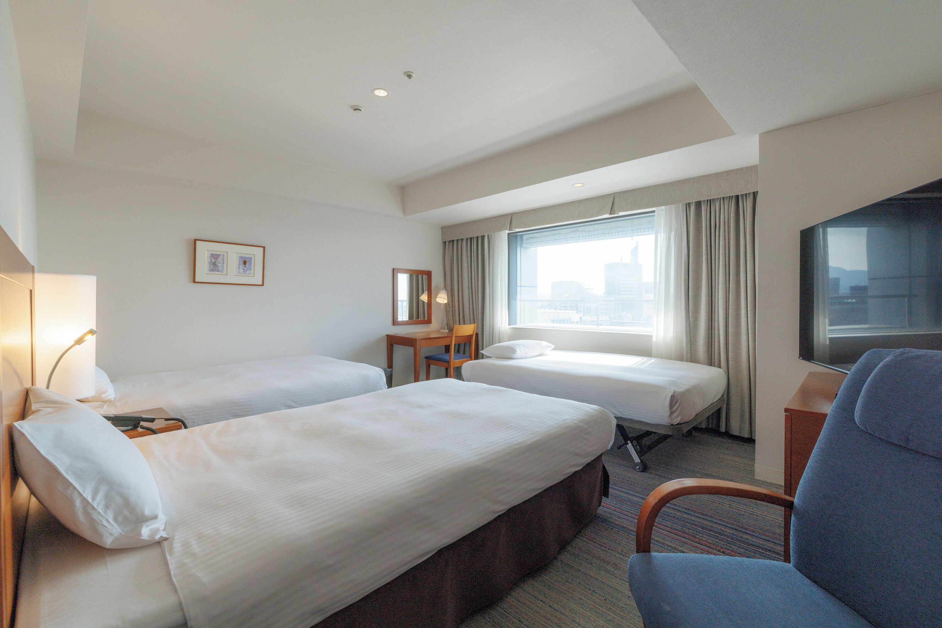 Ein Hotelzimmer mit zwei Einzelbetten, einem Schreibtisch, einem Stuhl, einem Fernseher und einem Vorhangfenster.