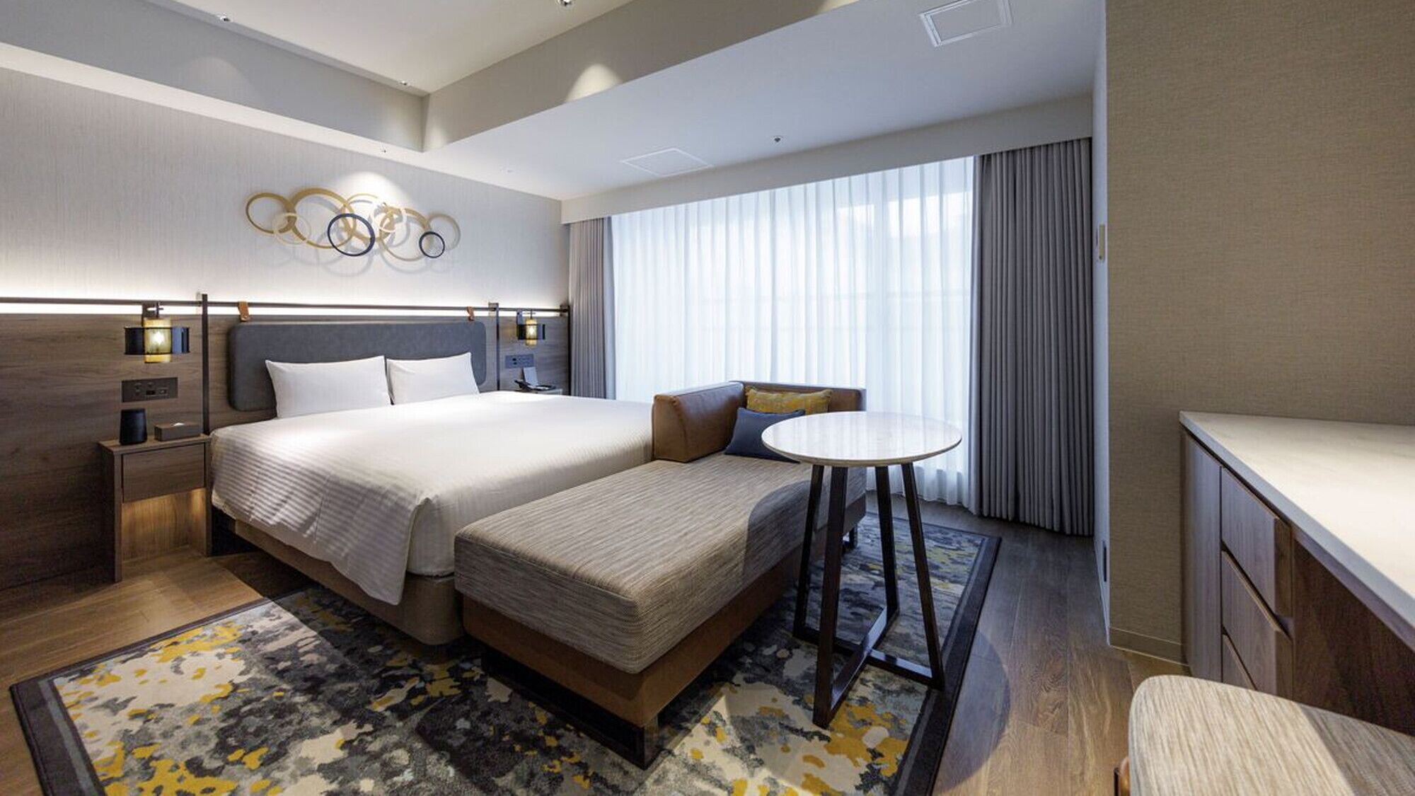 Une chambre d’hôtel avec un lit double, un canapé, une petite table, un bureau et une grande fenêtre avec des rideaux.