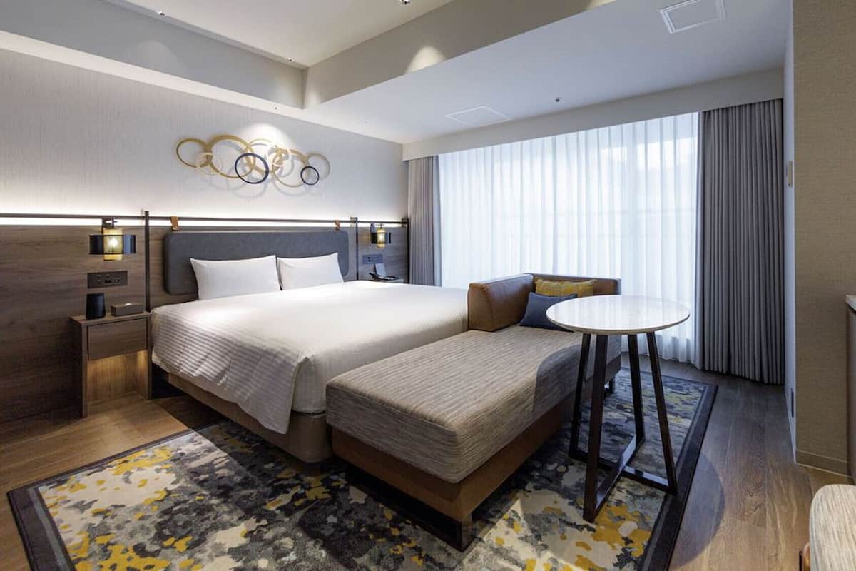Une chambre d’hôtel avec un lit double, un canapé, une petite table, un bureau et une grande fenêtre avec des rideaux.