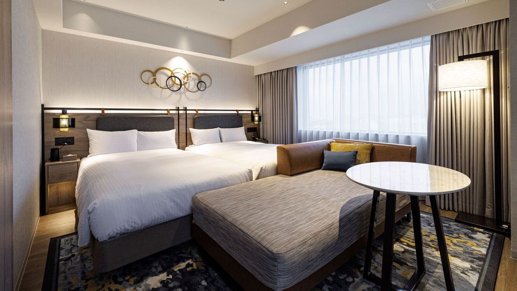 Une chambre d’hôtel avec deux lits, un canapé, une table ronde et une grande fenêtre avec des rideaux.